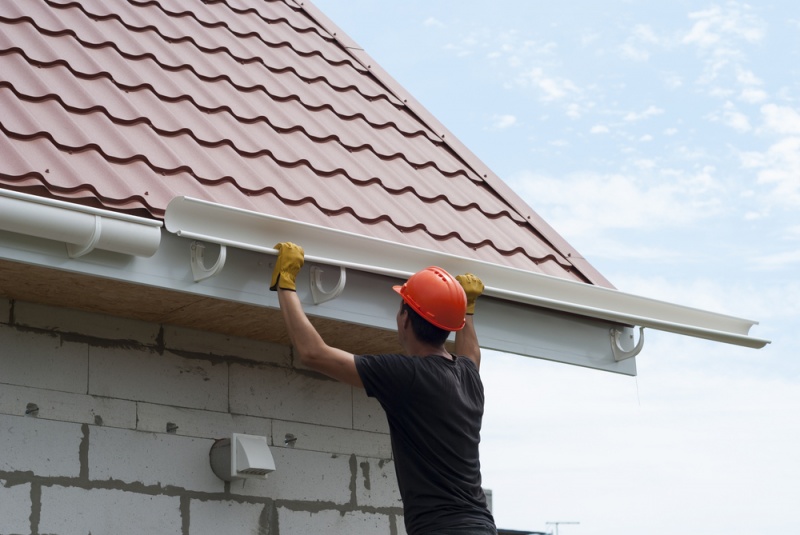 A close up shot of a worker in a black shirt and orange construction hat installing a set of white gutters on a sloped roof.